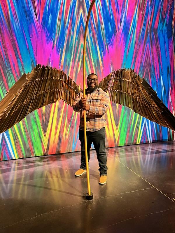 Malik Lockhart standing with sculptural wings against a vivid multicolor light backdrop